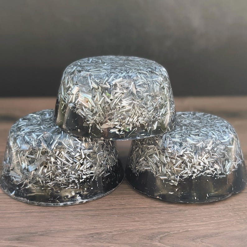 May include: Three black, dome-shaped objects with a silver, glitter-like material embedded throughout. The objects are stacked on top of each other, with the bottom two resting on a wooden surface. The background is a dark grey.