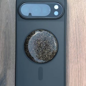 May include: A black phone case with a circular, textured design in the center. The case has a camera cutout with lenses and a flash. The circular design appears to be made of a dark, organic material.