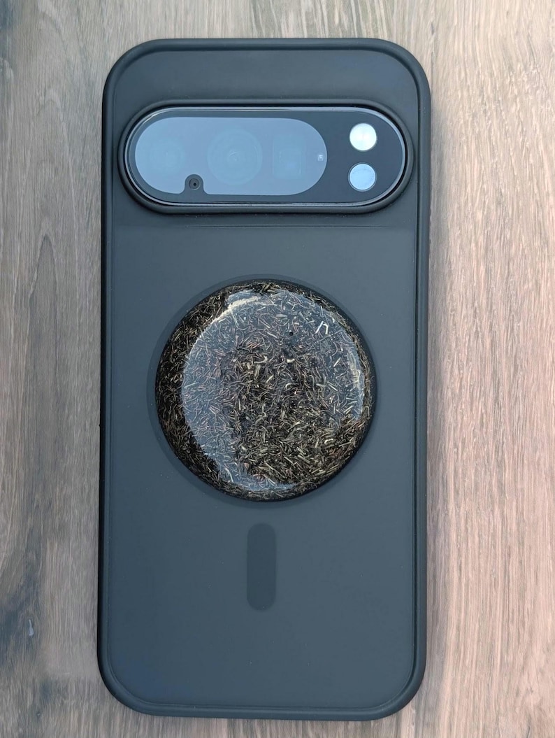 May include: A black phone case with a circular, textured design in the center. The case has a camera cutout with lenses and a flash. The circular design appears to be made of a dark, organic material.