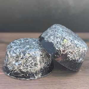 May include: Two black resin objects with embedded silver-coloured metallic flakes. One is a rounded dome shape, and the other is a rectangular shape. The objects are on a wooden surface.