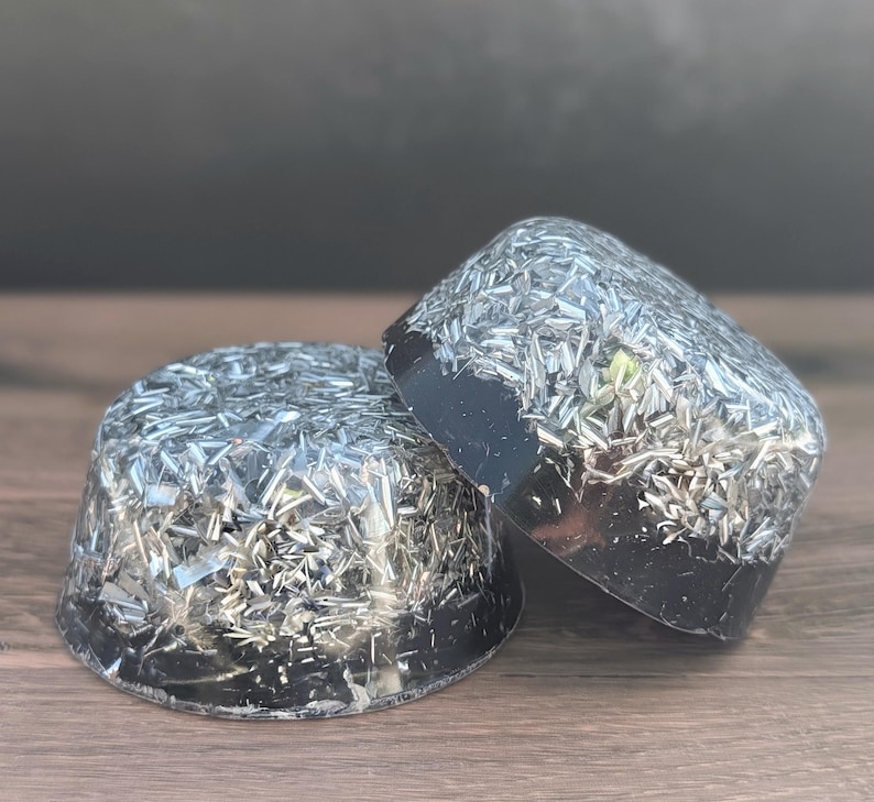 May include: Two black resin objects with embedded silver-coloured metallic flakes. One is a rounded dome shape, and the other is a rectangular shape. The objects are on a wooden surface.