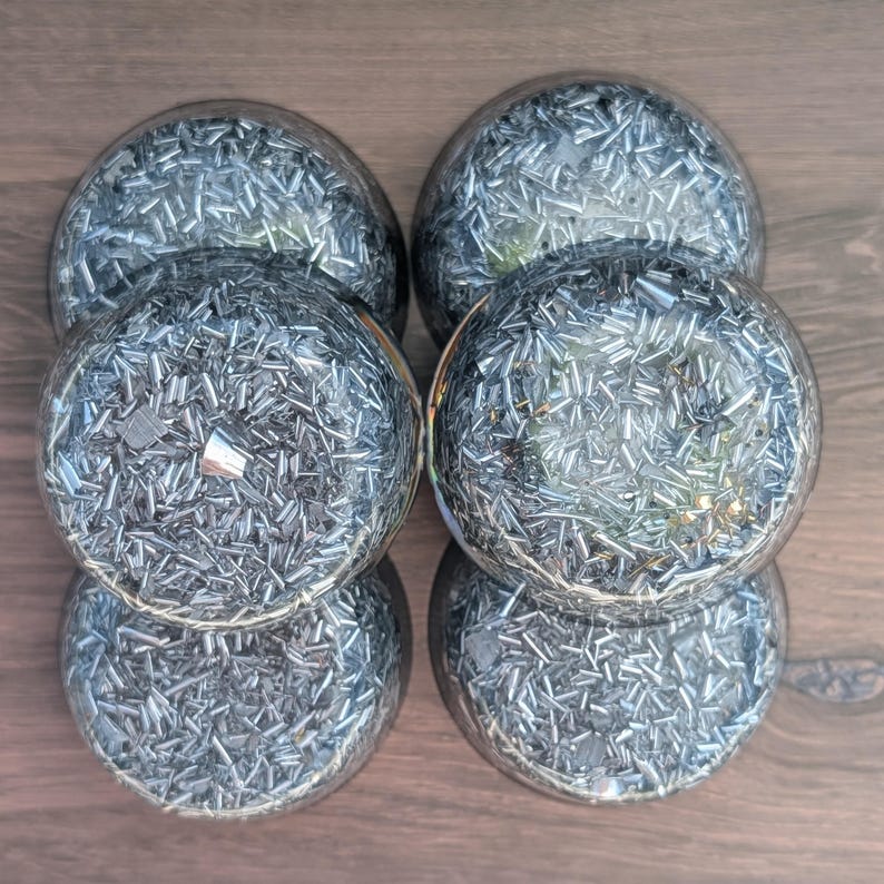 May include: Six clear glass spheres filled with silver-coloured, small, metallic pieces. The spheres are arranged in a circular pattern on a dark wood surface. The metallic pieces reflect light, creating a shimmering effect.