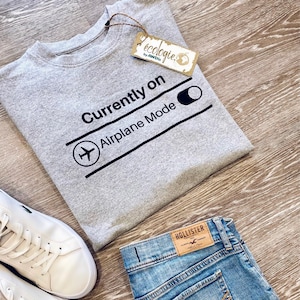 May include: A gray sweatshirt with the text "Currently on Airplane Mode" and a graphic of an airplane. The sweatshirt is paired with denim shorts and white sneakers. A tag from "&eacute;cologie by AWDis" is attached.