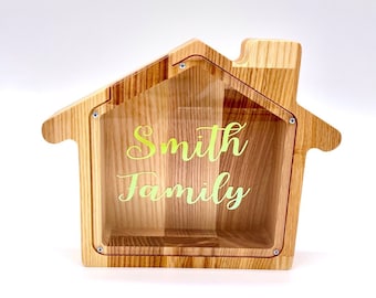 Puede incluir: Una alcancía de madera con forma de casa con un frente de plástico transparente. El frente tiene una calcomanía de vinilo amarilla y verde que dice "Smith Family".