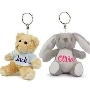 Könnte beinhalten: Zwei Plüsch-Schlüsselanhänger, einer ein brauner Teddybär, der ein blaues Shirt mit dem Namen "Jack" in Blau bestickt trägt, und der andere ein grauer Hase mit dem Namen "Olivia" in Pink bestickt.