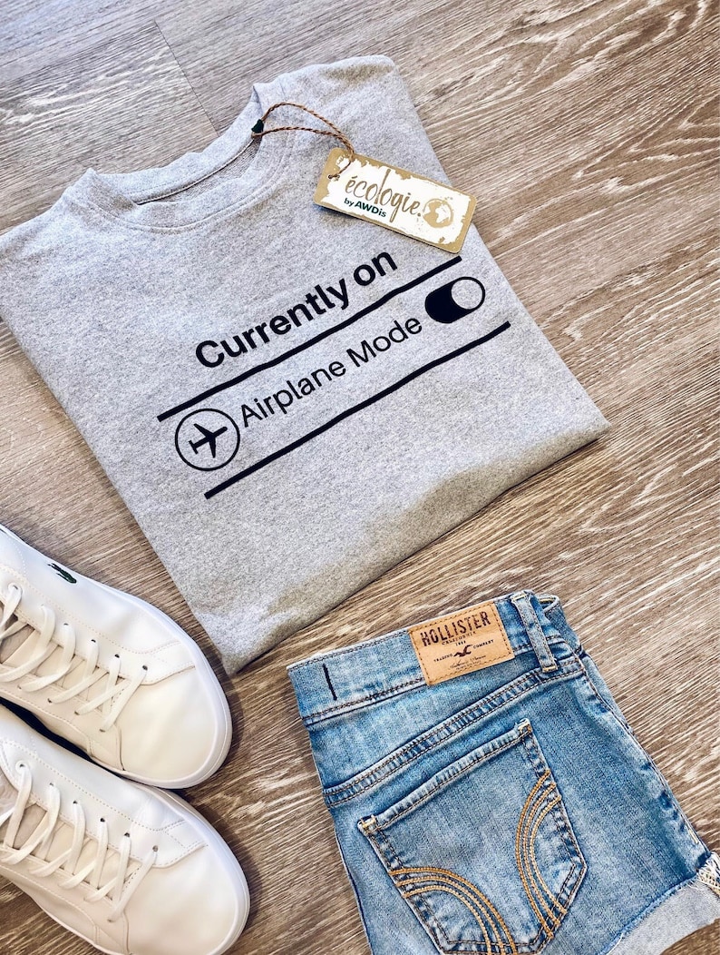 May include: A gray sweatshirt with the text "Currently on Airplane Mode" and a graphic of an airplane. The sweatshirt is paired with denim shorts and white sneakers. A tag from "&eacute;cologie by AWDis" is attached.