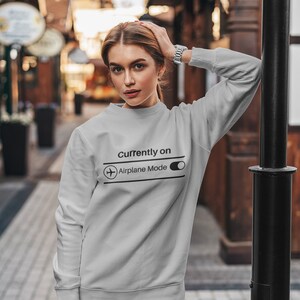 May include: A light gray sweatshirt with the text "Currently On Airplane Mode" and an airplane graphic. The sweatshirt is long-sleeved and is being worn by a person. The person is wearing blue jeans and a silver watch.