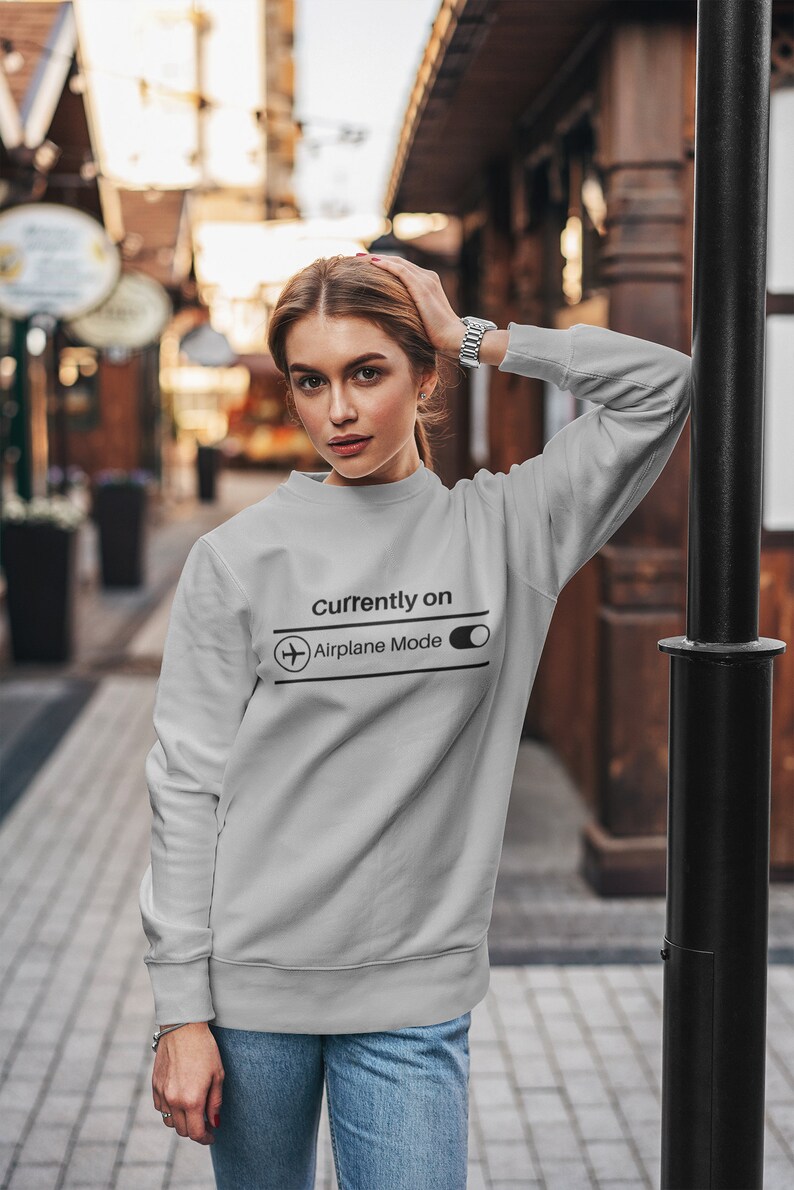 May include: A light gray sweatshirt with the text "Currently On Airplane Mode" and an airplane graphic. The sweatshirt is long-sleeved and is being worn by a person. The person is wearing blue jeans and a silver watch.