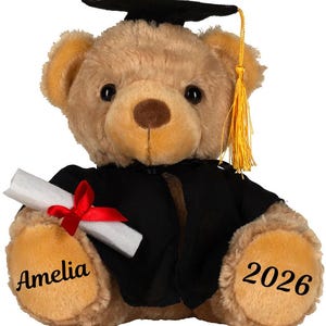 May include: A soft teddy bear dressed in a black graduation gown and mortarboard hat with a gold tassel. The bear holds a scroll tied with a red ribbon. The name "Amelia" and the year "2026" are embroidered on the bear's paws.