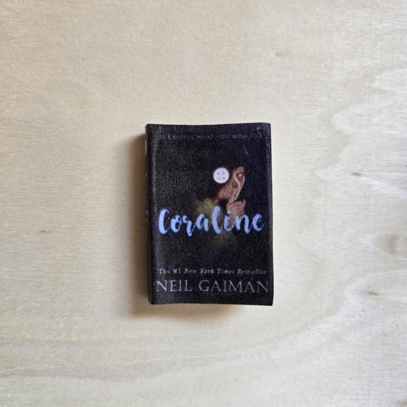 Op de afbeelding: Een zwart paperback boek met een blauwe en witte illustratie van een meisje met een knoopoog. De titel "Coraline" is geschreven in blauwe cursieve letters. Het boek is van Neil Gaiman en is een New York Times bestseller.
