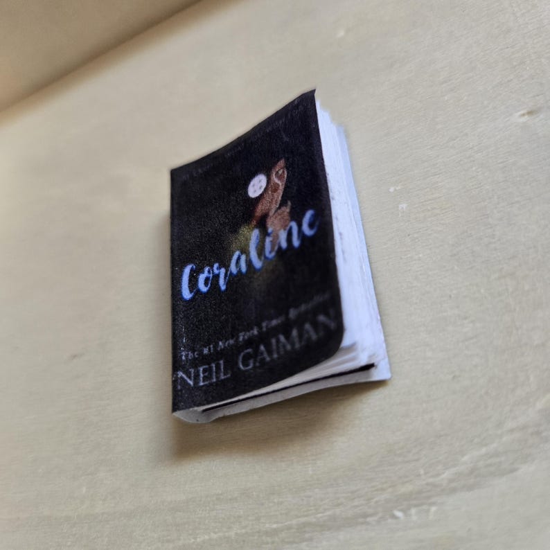 Op de afbeelding: Een miniatuur paperback boek met een zwarte omslag met een illustratie van een uil en de titel "Coraline" in blauwe letters. De auteur, Neil Gaiman, staat onder de titel gedrukt.
