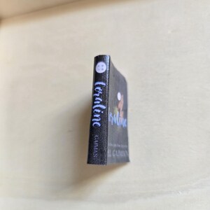 Op de afbeelding: Een zwart paperback boek getiteld "Coraline" van Neil Gaiman. De omslag van het boek toont een silhouet van een meisje met een kat op haar schouder.