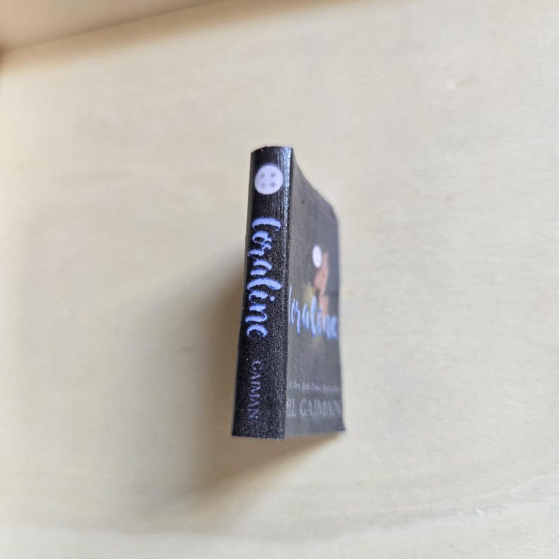 Op de afbeelding: Een zwart paperback boek getiteld "Coraline" van Neil Gaiman. De omslag van het boek toont een silhouet van een meisje met een kat op haar schouder.