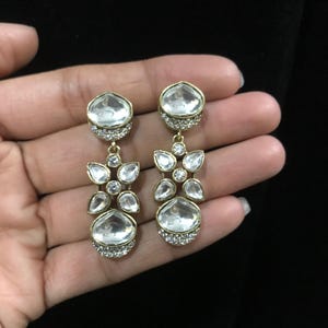 Pode incluir: Brincos pendurados dourados com pedras claras e facetadas. Os brincos apresentam um conjunto de pedras claras em um design floral, com uma pedra maior em forma de gota na parte inferior. Os brincos s&atilde;o segurados em uma m&atilde;o contra um fundo preto.