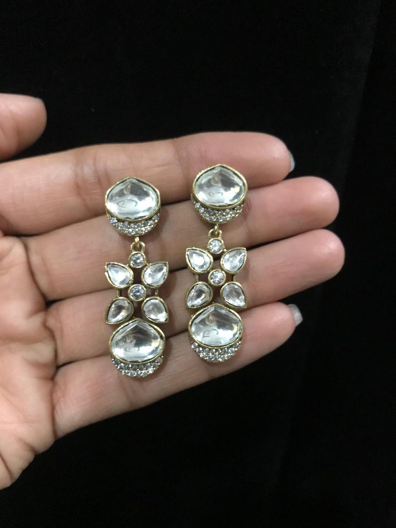 Pode incluir: Brincos pendurados dourados com pedras claras e facetadas. Os brincos apresentam um conjunto de pedras claras em um design floral, com uma pedra maior em forma de gota na parte inferior. Os brincos s&atilde;o segurados em uma m&atilde;o contra um fundo preto.
