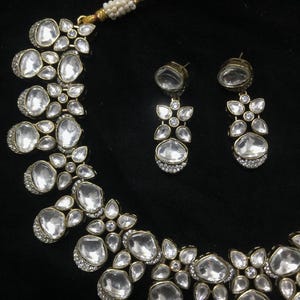 Pode incluir: Um conjunto de colar e brincos a condizer em tom dourado. As joias apresentam pedras grandes, claras, ovais e em forma de gota, acentuadas com pedras claras mais pequenas. O conjunto est&aacute; sobre um fundo preto.