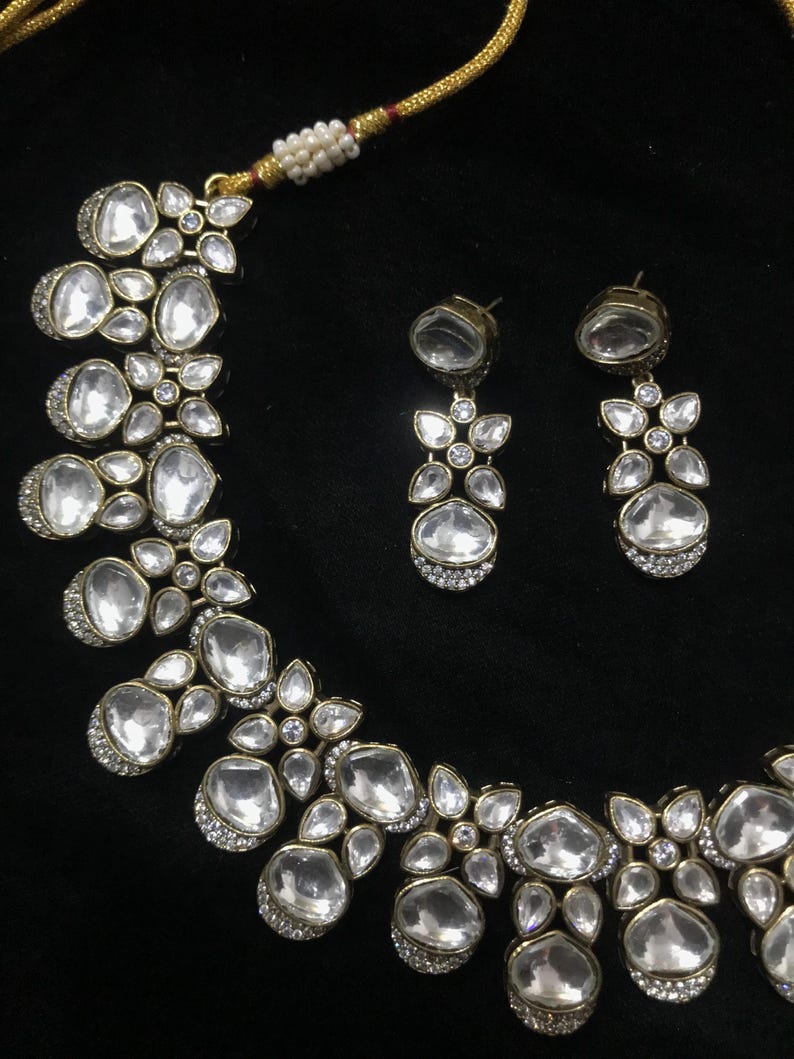 Pode incluir: Um conjunto de colar e brincos a condizer em tom dourado. As joias apresentam pedras grandes, claras, ovais e em forma de gota, acentuadas com pedras claras mais pequenas. O conjunto est&aacute; sobre um fundo preto.
