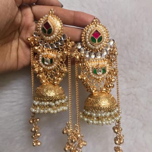 Op de afbeelding: Goudkleurige oorbellen met ingewikkelde details. Elke oorbel heeft een bloemmotief met rode en groene accenten, omringd door parels en heldere stenen. Hangende kettingen en kleine gouden kralen voegen textuur toe.