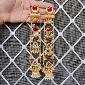 May include: Gold-toned earrings with red gemstone accents and dangling gold beads. The earrings feature a floral design with pink and green details, and end with a bell-shaped bottom. The earrings are held by a hand.