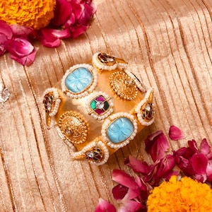 Op de afbeelding: Een goudkleurige armband met ingewikkelde details, met ovale turquoise stenen omringd door kleine parels. Extra gouden accenten zijn onder meer kleinere, sierlijke cirkelvormige ontwerpen en juweelkleurige versieringen. De armband ligt op een gouden stoffen achtergrond.