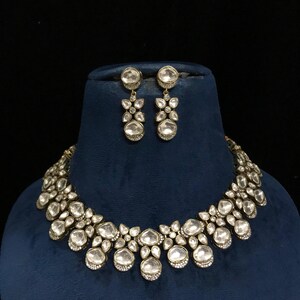 Pode incluir: Um conjunto de colar e brincos a condizer em tom prateado. As joias apresentam pedras grandes, claras, redondas e em forma de gota, engastadas num design floral. O colar e os brincos s&atilde;o exibidos num busto de veludo azul sobre um fundo preto.