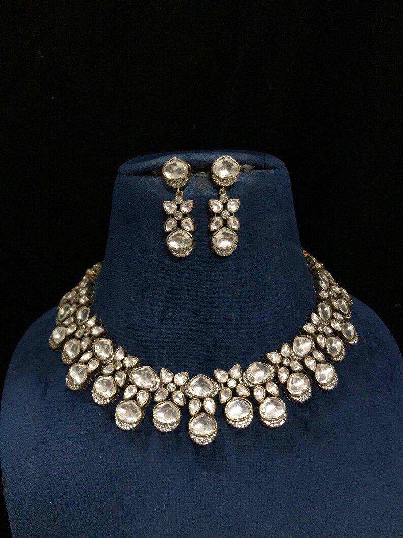 Pode incluir: Um conjunto de colar e brincos a condizer em tom prateado. As joias apresentam pedras grandes, claras, redondas e em forma de gota, engastadas num design floral. O colar e os brincos s&atilde;o exibidos num busto de veludo azul sobre um fundo preto.