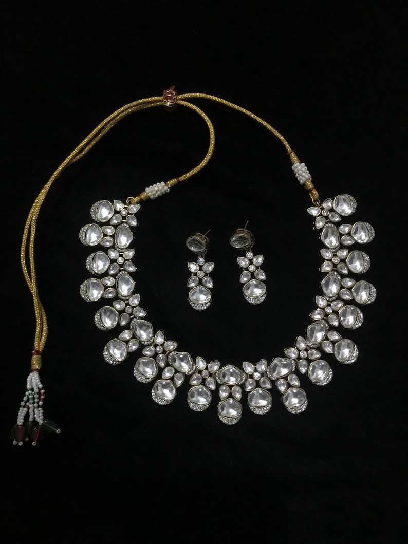 Pode incluir: Um conjunto de colar e brincos prateados. O colar apresenta pedras grandes e claras em forma oval, rodeadas por pedras mais pequenas e claras. Os brincos espelham o design. O conjunto &eacute; exibido sobre um fundo preto.