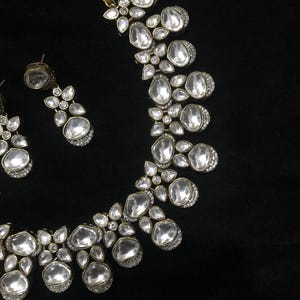 Pode incluir: Um conjunto de colar e brincos prateados. As joias apresentam pedras grandes e claras em forma oval, rodeadas por pequenos detalhes brilhantes. O colar e os brincos est&atilde;o sobre um fundo preto.