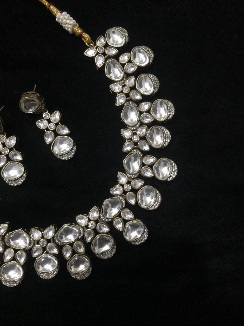 Pode incluir: Um conjunto de colar e brincos prateados. As joias apresentam pedras grandes e claras em forma oval, rodeadas por pequenos detalhes brilhantes. O colar e os brincos est&atilde;o sobre um fundo preto.