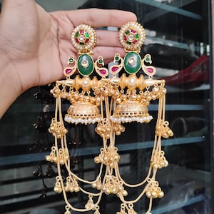 Op de afbeelding: Goudkleurige oorbellen met ingewikkelde details. Elke oorbel heeft een cirkelvormige bovenkant met rode, groene en witte accenten, een groene ovale steen en een pauwenmotief. Hangende kettingen met kleine gouden belletjes maken het ontwerp compleet.