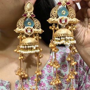 Puede incluir: Pendientes dorados con diseño de araña de luces. Cada pendiente presenta una piedra preciosa azul claro, detalles rojos y pequeñas perlas blancas. Los pendientes tienen un elemento en forma de campana y múltiples cadenas con pequeñas cuentas doradas.