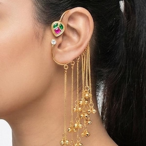 May include: Gold-toned ear cuff featuring a heart-shaped design with green and pink accents, a dangling chain, and small gold beads. The ear cuff also has a small, clear gemstone. This piece is a unique jewelry accessory.