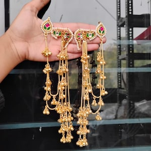 Op de afbeelding: Een paar goudkleurige oorbellen met ingewikkelde details. Elke oorbel heeft een druppelvormige bovenkant met rode, groene en witte accenten en een waterval van hangende gouden kralen. De oorbellen worden door een hand omhoog gehouden.