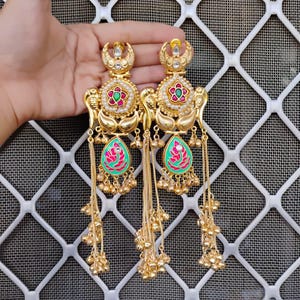 Op de afbeelding: Goudkleurige oorhangers met ingewikkelde bloemen- en druppelontwerpen. De oorbellen hebben rode, groene en witte accenten, met bungelende kettingen die eindigen in kleine gouden kralen. De oorbellen zijn sierlijk en gedetailleerd.