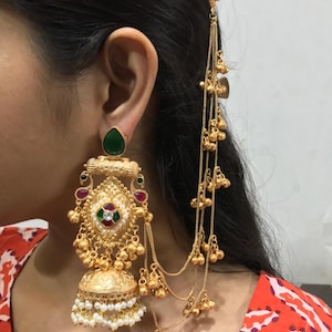 May include: Gold-toned earring with a teardrop-shaped green stone, intricate filigree, and dangling chains with small bells. The earring features red, green, and white accents, and a bell-shaped bottom with white pearls.