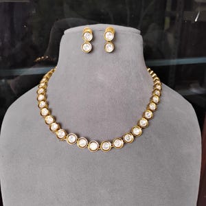 Peut inclure: Un ensemble collier et boucles d'oreilles assorties de couleur or. Le collier et les boucles d'oreilles présentent des pierres rondes et claires serties dans du métal doré. Les bijoux sont exposés sur un mannequin gris.