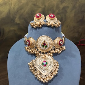 Op de afbeelding: Goudkleurige sieradenset met een ketting en bijpassende oorbellen. De ketting heeft een hartvormige hanger met rode, groene en witte accenten. De oorbellen en ketting hebben een bloemenontwerp met rode edelstenen.