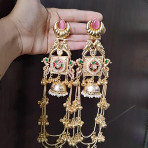 Op de afbeelding: Een paar goudkleurige oorbellen met een roze edelsteen aan de bovenkant. De oorbellen hebben ingewikkelde ontwerpen, waaronder vierkante elementen met rode, groene en witte accenten, en bungelende kettingen met kleine gouden kralen. De oorbellen zijn sierlijk en gedetailleerd.