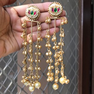 May include: Gold-toned dangle earrings with a floral design. Each earring features a round top with a pearl border, red and green accents, and cascading chains of small beads and larger pearls. The earrings are designed for pierced ears.