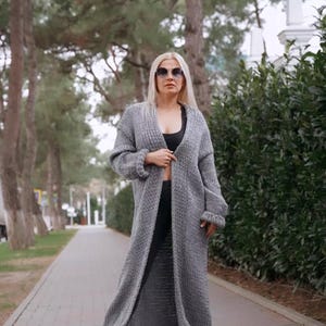 May include: A long, gray knitted cardigan with a V-neck and long sleeves. The cardigan is open in the front and the person wearing it is standing on a paved path.