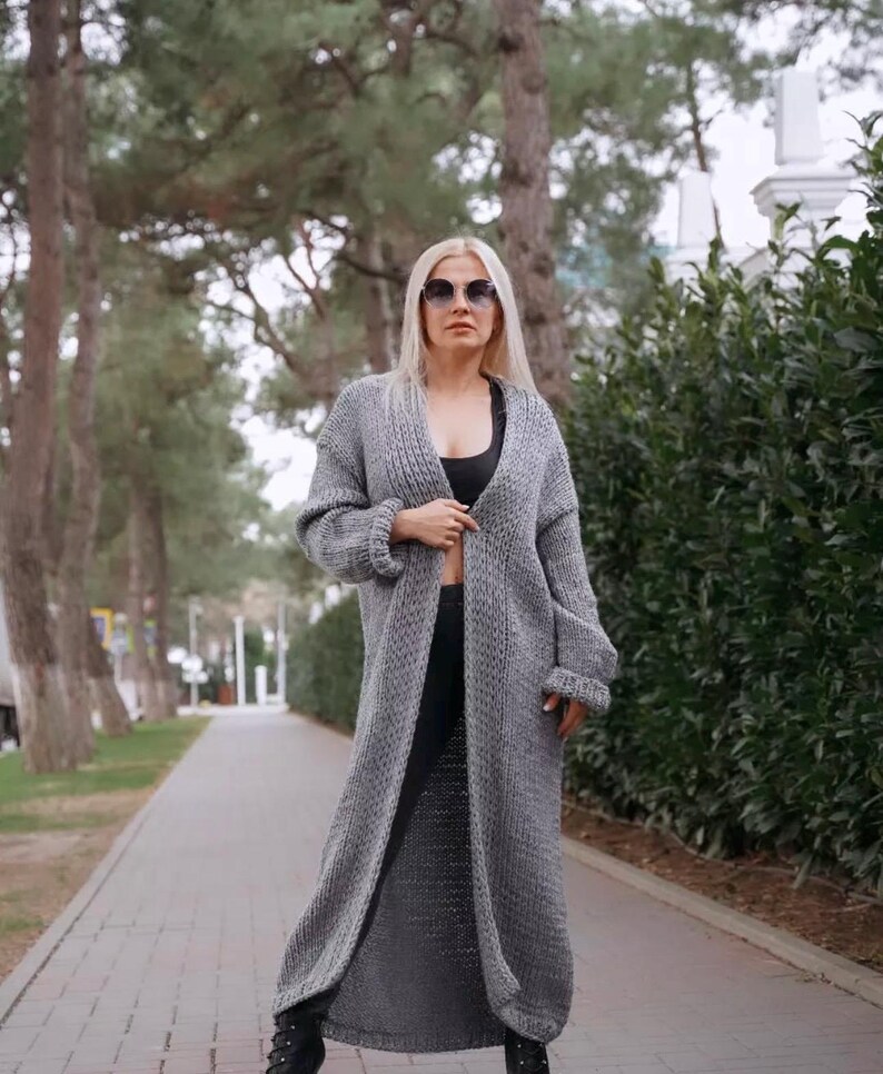 May include: A long, gray knitted cardigan with a V-neck and long sleeves. The cardigan is open in the front and the person wearing it is standing on a paved path.