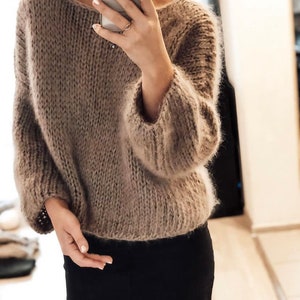 Könnte beinhalten: Ein übergroßer Strickpullover in Taupe mit weitem Ausschnitt und langen Ärmeln. Der Pullover hat eine flauschige Textur und wird mit einem schwarzen Rock kombiniert. Die Person auf dem Bild hält ein Telefon.