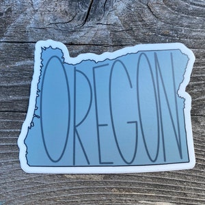 May include: A gray vinyl sticker of the state of Oregon with the word "OREGON" written in a stylized font inside the outline of the state.