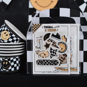 Puede incluir: Una caja de regalo de fiesta con un patrón de cuadros blanco y negro. La caja contiene un conjunto de tatuajes temporales con una patineta, rayos, estrellas y caras sonrientes. La caja también tiene el texto "Young, Wild & Three" y "Tattoos".