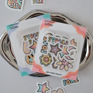 May include: Two packs of temporary tattoos with a variety of designs, including stars, hearts, smiley faces, and the words "groovy" and "love". The packs are on a silver tray and have a pink, orange, and blue color scheme.