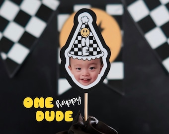 May include: A black cupcake with chocolate frosting and a yellow and black checkered birthday hat with a smiling face and the number "ONE" on it. The cupcake is decorated with the words "ONE happy DUDE" in yellow letters.