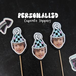 May include: Three personalized cupcake toppers featuring a child's photo, each with a checkered party hat design. The toppers are on wooden sticks, surrounded by decorative stickers with a black background. Text on the image reads "PERSONALIZED Cupcake Toppers."