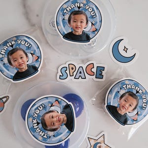 Puede incluir: Tres contenedores de plástico transparente con pegatinas de temática espacial azul y blanca con la foto de un niño y el texto "¡Gracias!" Las pegatinas también incluyen estrellas, una luna creciente y un planeta. También hay una pegatina que dice "ESPACIO" en letras de arcoíris.