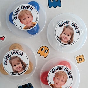Peut inclure: Quatre contenants en plastique transparent contenant des toupies à fidgets colorées. Chaque toupie est décorée d'un autocollant rond avec la photo d'un enfant et le texte "GAME OVER" avec un design pixélisé. Les autocollants indiquent également "MERCI D'AVOIR JOUÉ !"