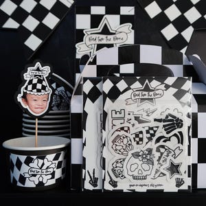 Puede incluir: Suministros para fiestas a cuadros en blanco y negro con el texto "Bad to the Bone" y gr&aacute;ficos de calaveras y huesos cruzados. Los suministros incluyen adornos para cupcakes, vasos y una caja de regalo con pegatinas.